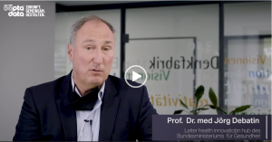 Videostandbild eines Mannes mittleren Alters im dunklen Sakko und hellblauem Hemd, der in die Kamera spricht; Ansteckmikrofon sichtbar. Im Hintergrund ein verglaster Besprechungsraum mit Beschriftungen auf der Scheibe. Links oben das Logo „50 Jahre opta data – Zukunft. Gemeinsam. Gestalten.“; in der Bauchbinde unten rechts stehen Name und Funktion (Leitung des health innovation hub des Bundesministeriums für Gesundheit). In der Mitte liegt ein Play-Symbol.