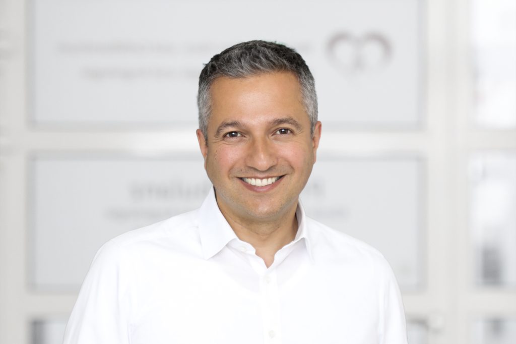 Porträt eines Mannes mittleren Alters mit kurzen, dunkelgrau melierten Haaren und freundlichem Lächeln. Er trägt ein weißes Hemd und blickt direkt in die Kamera. Der Oberkörper ist frontal zu sehen. Im hellen, unscharfen Hintergrund hängen mehrere gerahmte Dokumente oder Urkunden an der Wand, was auf eine Büro- oder Praxisumgebung hindeutet.