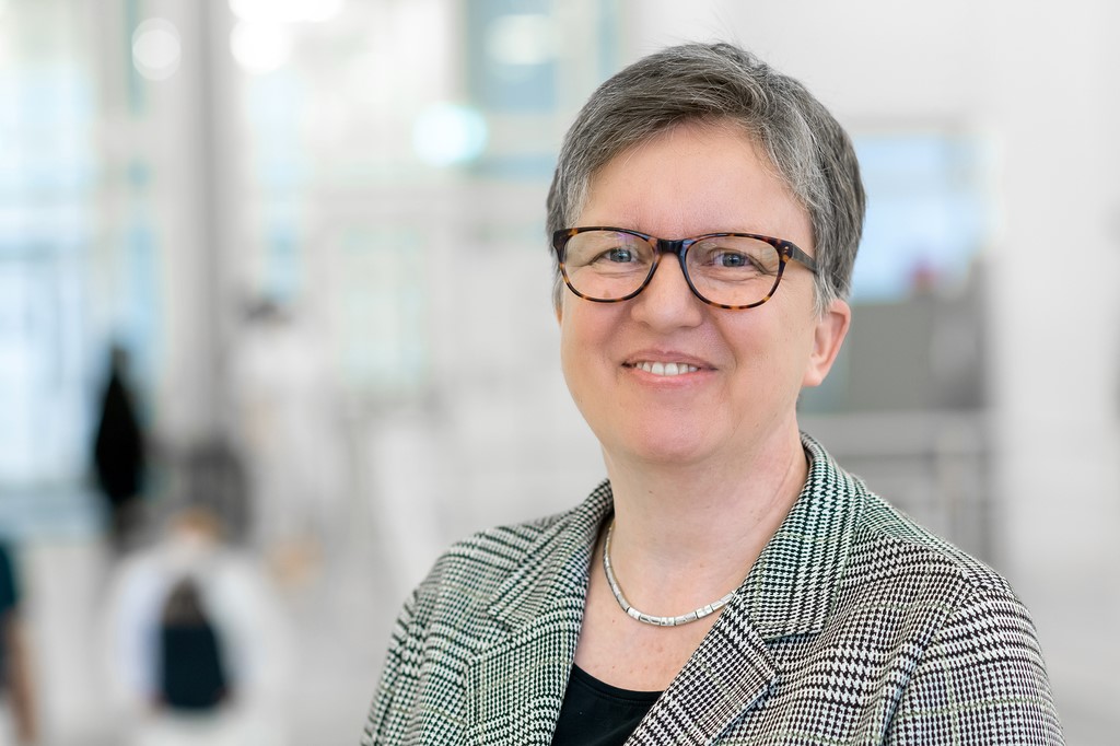 Porträt einer lächelnden Person mit kurzem grauem Haar und Brille. Sie trägt ein dunkles Oberteil sowie einen grau gemusterten Blazer und blickt direkt in die Kamera. Im Hintergrund sind helle, unscharfe Innenräume mit großen Fenstern zu erkennen, wodurch ein freundlicher und professioneller Eindruck entsteht.