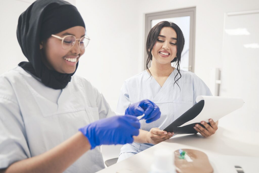 Zwei junge Frauen in medizinischer Kleidung sitzen in einem hellen Raum. Eine von ihnen trägt Handschuhe und arbeitet konzentriert, während die andere lächelnd ein Klemmbrett mit Unterlagen hält.