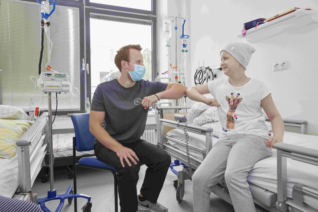 In einem hellen Krankenhauszimmer sitzen sich ein maskentragender Erwachsener auf einem Stuhl und ein Kind mit Mütze auf dem Klinikbett gegenüber und geben sich lächelnd einen Ellenbogen-Gruß. Neben dem Bett stehen Infusionsständer und medizinische Geräte; auf dem T-Shirt des Kindes ist eine bunte Giraffe. Unterstützende, zugewandte Szene.