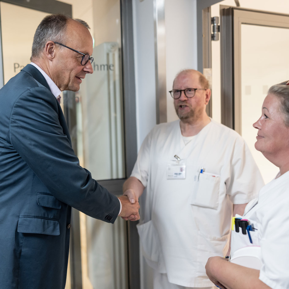 Bundeskanzler Friedrich Merz schüttelt einem Krankenhausmitarbeiter in weißer Arbeitskleidung die Hand. Neben ihnen steht eine weitere Mitarbeiterin im weißen Kittel, die den Händedruck beobachtet.