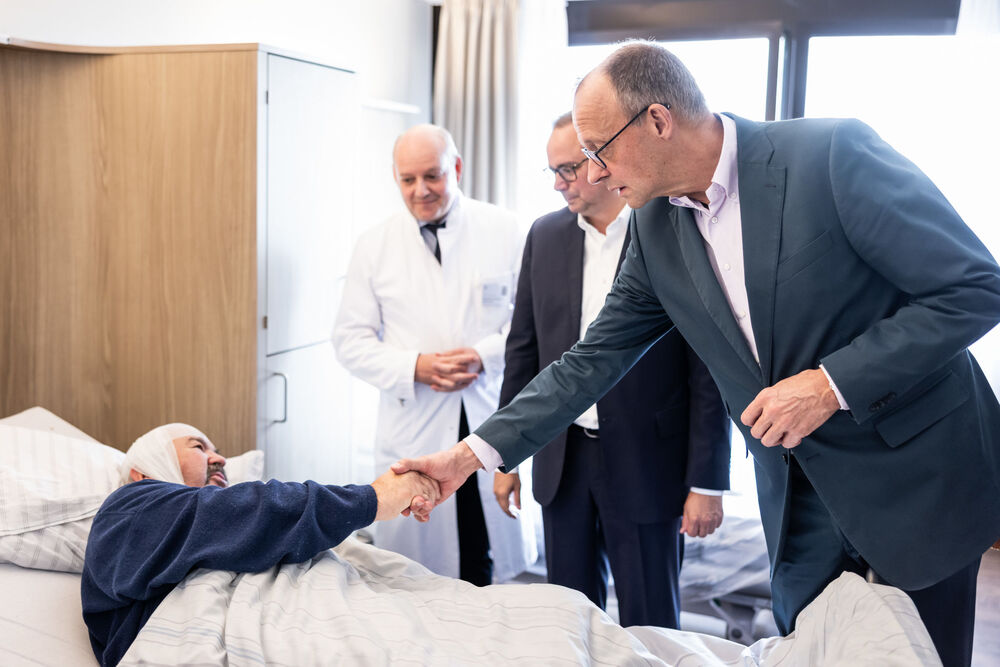 Bundeskanzler Friedrich Merz schüttelt einem Patienten im Krankenhausbett die Hand. Der Patient liegt zugedeckt und trägt einen Kopfverband. Im Hintergrund stehen der Ärztliche Direktor des Alfried Krupp Krankenhaus Dr. Stephan Buse und Oberbürgermeister Thomas Kufen beobachten die Begegnung.