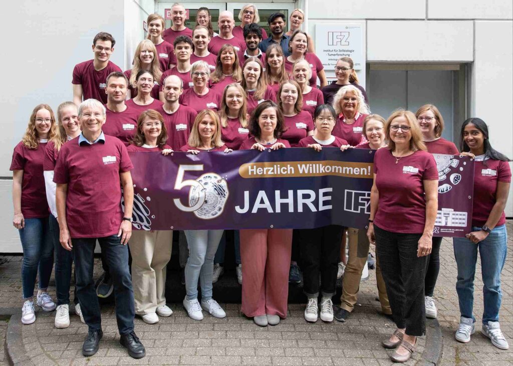 Gruppenfoto von Mitarbeitenden des Instituts für Zellbiologie, die in bordeauxfarbenen T-Shirts vor dem Gebäude stehen und ein Banner mit der Aufschrift ‚50 Jahre IFZ – Herzlich Willkommen!‘ halten.