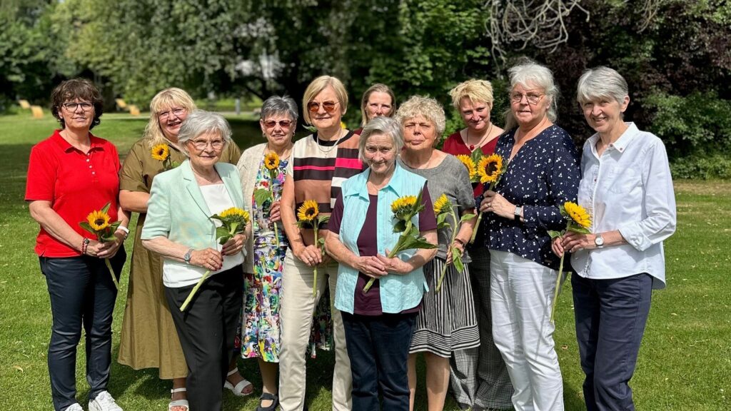Eine Gruppe von elf Frauen steht auf einer Wiese im Sonnenschein und hält jeweils eine Sonnenblume in der Hand. Sie lächeln in die Kamera; im Hintergrund sind Bäume und Grünflächen zu sehen.