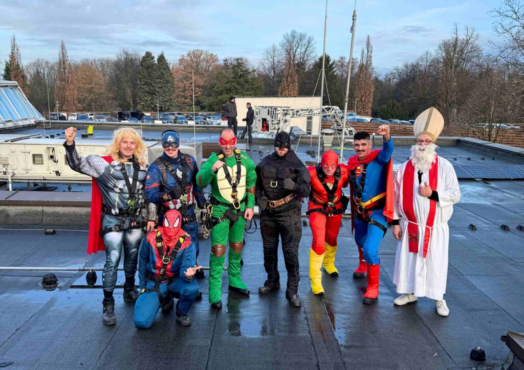 Auf einem flachen Gebäudedach stehen neun verkleidete Personen nebeneinander und posieren fröhlich für die Kamera. Acht von ihnen tragen bunte Superheldenkostüme, darunter Figuren mit roten, blauen, grünen und schwarzen Ganzkörperanzügen, Masken und Umhängen. Eine Person ist als Nikolaus in weißer Robe mit rotem Schal, weißem Bart und hoher Bischofsmütze verkleidet. Einige tragen Sicherungsgurte, was darauf hinweist, dass sie sich auf dem Dach gesichert haben. Der Boden ist leicht feucht. Im Hintergrund sind technische Dachaufbauten, mehrere parkende Autos und winterliche Bäume zu sehen. Die Gruppe wirkt ausgelassen und hält teilweise die Fäuste siegreich in die Höhe.