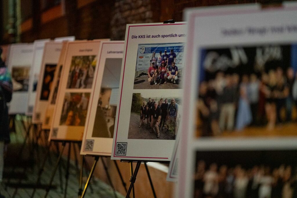 Mehrere großformatige Plakate stehen dicht nebeneinander auf schwarzen Metallstativen in einer leicht schrägen Reihe. Sie zeigen verschiedene Fotos und kurze Überschriften, die auf schulische oder gemeinschaftliche Aktivitäten hinweisen. Im Vordergrund ist ein Plakat gut erkennbar, darauf zwei Gruppenfotos von sportlich gekleideten Personen; oben steht der Titel „Die KKS ist auch sportlich unter…“. Jedes Plakat trägt unten einen QR-Code. Die Szene ist in warmes, leicht gedämpftes Licht getaucht, vermutlich am Abend oder in einem Innenhof. Der Hintergrund bleibt unscharf, lässt aber eine Mauer und