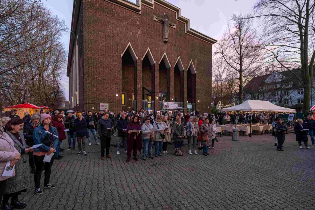 Auf einem offenen Platz vor einer großen Backsteinkirche mit markantem Eingangsportal und Kreuz versammelt sich eine größere Gruppe von Menschen. Es ist früher Abend, der Himmel ist blass und die Szene wirkt winterlich; viele Personen tragen Jacken, Schals und Mützen. Die Menschen stehen locker verteilt, einige halten Blätter in den Händen, als würden sie an einer gemeinschaftlichen Aktion wie einem Liedersingen oder einer Andacht teilnehmen. Links und rechts am Rand stehen kleine Markt- oder Infostände mit Zeltdächern und Lichterketten. Im Hintergrund sind kahle Bäume und Wohnhäuser zu sehen. Die Stimmung wirkt ruhig und konzentriert.