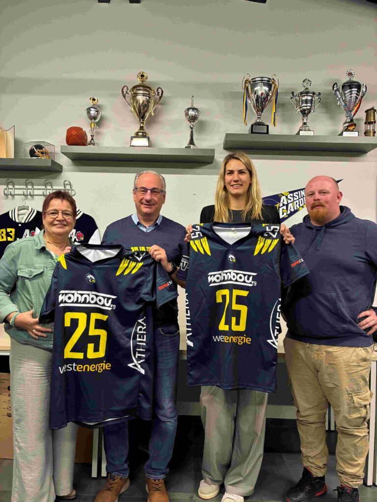 Vier Personen stehen nebeneinander in einem Raum vor einer Wand mit zahlreichen Pokalen und Trophäen auf Regalen. Alle vier blicken lächelnd in die Kamera. Die beiden Personen in der Mitte halten jeweils ein dunkelblaues American-Football-Trikot mit der gelben Nummer 25 und mehreren Sponsorenlogos in den Händen. Links steht eine Person mit kurzem Haar und Brille, rechts eine Person mit Bart und Kapuzenpullover. Die Szene wirkt wie eine Übergabe oder Präsentation der Trikots.