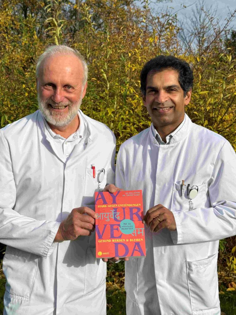 Zwei Personen in weißen Laborkitteln stehen dicht nebeneinander im Freien vor herbstlich gelb-grünem Buschwerk. Beide lächeln freundlich in die Kamera. Gemeinsam halten sie ein bunt gestaltetes Buch mit roter und orangefarbener Titelseite, auf der groß das Wort „Ayurveda“ sowie weitere Schriftzüge in verschiedenen Sprachen und ein blauer Sticker zu sehen sind.
