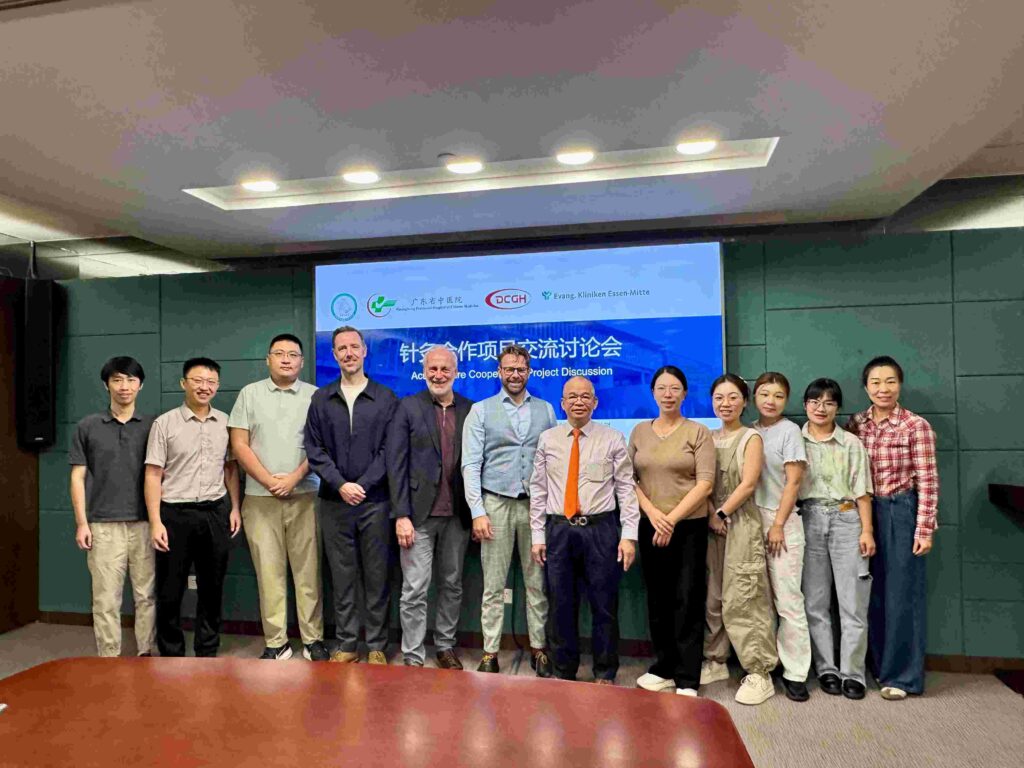 Eine Gruppe von dreizehn Personen steht in einem Konferenzraum nebeneinander und blickt zur Kamera. Die Gruppe besteht aus mehreren Männern und Frauen verschiedener Altersgruppen. Sie tragen überwiegend Business- oder Business-Casual-Kleidung. Hinter ihnen hängt eine große Präsentationsfolie mit Logos und zweisprachigem Titel zur deutsch-chinesischen Projektzusammenarbeit in der Medizin beziehungsweise Naturheilkunde. Der Raum hat grüne Wandverkleidungen und eine helle Deckenbeleuchtung. Im Vordergrund ist ein großer Holztisch angeschnitten.