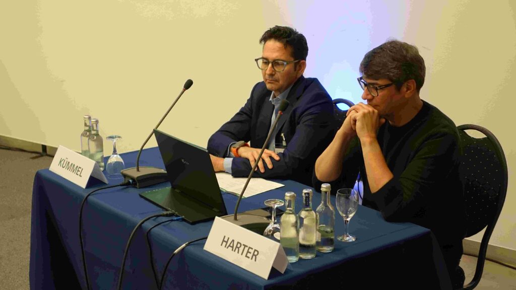 An einem mit dunkelblauem Tuch bedeckten Konferenztisch sitzen zwei Männer nebeneinander. Beide tragen Brillen und blicken konzentriert nach vorn auf einen Laptop. Links sitzt ein Mann im dunkelblauen Sakko, vor ihm steht ein Namensschild mit der Aufschrift „Kümmel“ sowie ein Mikrofon, zwei Wasserflaschen und ein leeres Glas. Rechts neben ihm sitzt ein Mann in dunklem Oberteil, vor ihm das Namensschild „Harter“, ebenfalls ein Mikrofon und mehrere Flaschen mit Wasser. Die beiden wirken fokussiert, als würden sie einen Vortrag halten oder eine Diskussion moderieren. Der Hintergrund ist schlicht und hell, ohne weitere Details.