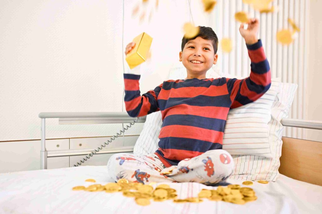 Ein kleiner Junge sitzt fröhlich auf einem Krankenhausbett. Er trägt ein langärmeliges Shirt mit roten und dunkelblauen Streifen sowie eine weiße Schlafhose mit bunten Comicmotiven. Mit beiden Armen wirft er goldene Schokomünzen in die Luft, sodass einige bereits um ihn herum auf der Bettdecke liegen. Hinter ihm sind weiße Kissen und ein neutraler Krankenhausraum zu sehen. Der Junge lächelt breit und wirkt ausgelassen und spielerisch.