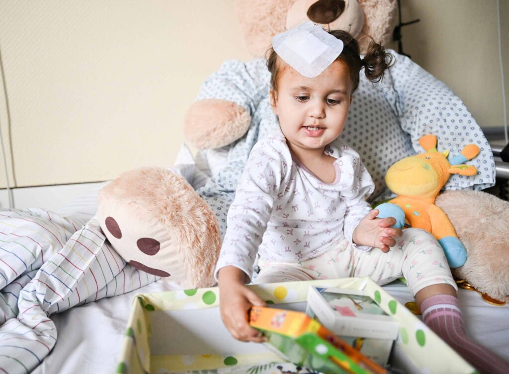 Ein kleines Kind sitzt in einem Krankenhausbett, umgeben von mehreren Stofftieren, darunter ein großer Teddy und eine bunte Stoffgiraffe. Das Kind trägt Schlafkleidung und hat ein medizinisches Pflaster auf der Stirn. Es greift fröhlich nach einem Spielzeug oder einer Schachtel in einer Geschenkbox auf dem Schoß. Die Bettdecke mit Streifen- und Punktmustern wirkt aufgeschlagen, und die Kuscheltiere sind eng um das Kind gruppiert, sodass eine warme, tröstliche Atmosphäre entsteht.