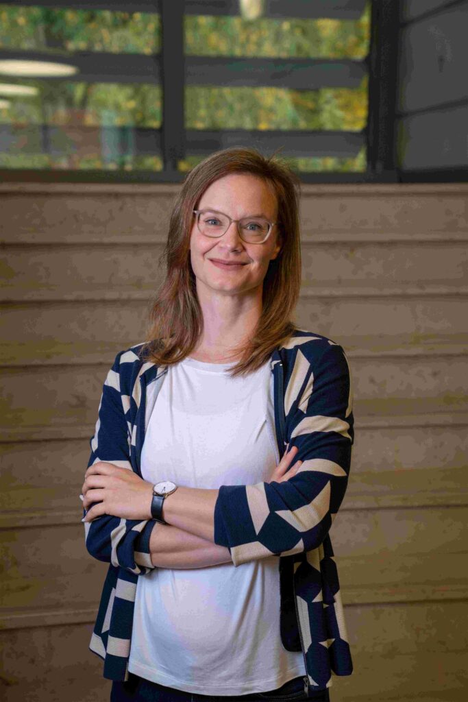 Porträt einer Frau mit schulterlangen, hellbraunen Haaren und Brille, die mit verschränkten Armen vor einer Holztreppe steht. Sie trägt ein weißes T-Shirt und eine dunkelblaue Strickjacke mit hellem geometrischem Muster. Die Frau blickt freundlich und selbstbewusst in die Kamera. Im Hintergrund sind Holzstufen und ein Geländer zu sehen, durch ein Fenster fällt Tageslicht. Die Umgebung wirkt modern und innenräumlich, etwa wie in einem Büro- oder Bildungsgebäude.