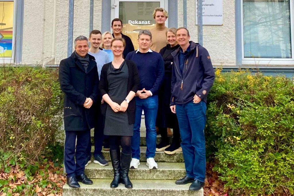 Neun Erwachsene stehen in mehreren Reihen auf einer kurzen Außentreppe vor dem Eingang eines hellen Gebäudes. In der Mitte befindet sich eine Glastür mit einem Schild darüber. Die Personen tragen überwiegend dunkle Alltags- und Freizeitkleidung, einige Jacken, eine Person vorne in schwarzen Stiefeln. Alle blicken in die Kamera und lächeln. Links und rechts der Treppe wachsen niedrige Sträucher, auf dem Boden liegen herbstliche Blätter. Die Szene wirkt wie ein Gruppenfoto vor einem Büro- oder Institutsgebäude.