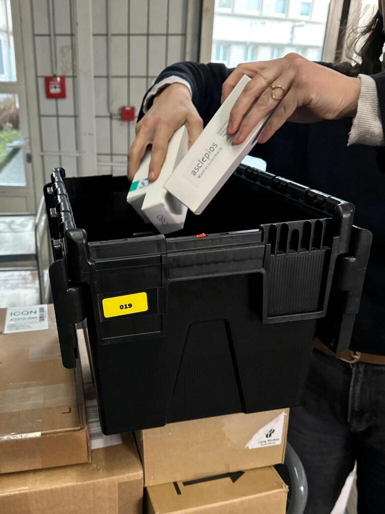 Zwei Hände legen mehrere weiße, schmale Medizinprodukt-Schachteln in eine offene schwarze Transportbox. Die Box steht auf gestapelten braunen Kartons und trägt einen gelben Aufkleber mit der Nummer „019“. Im Hintergrund ist ein heller Innenraum mit Fenstern und gefliesten Wänden zu sehen. Die Szene zeigt das Verpacken von medizinischem Material für den Versand.