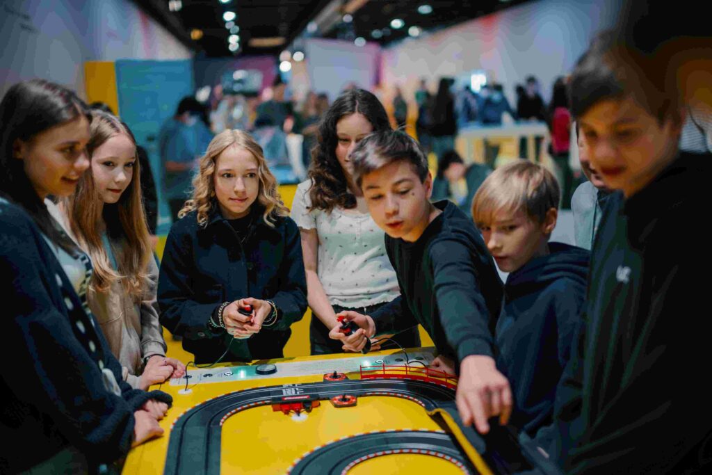 Mehrere Kinder stehen dicht um einen gelben Tisch und spielen gemeinsam mit einer Autorennbahn. In der Mitte verläuft eine schwarze Rennstrecke mit Kurven, darauf fahren kleine rote Fahrzeuge. Einige Kinder halten Handregler in den Händen, ein Junge beugt sich nach vorne und zeigt auf die Bahn. Alle blicken konzentriert auf das Spiel. Im Hintergrund ist ein heller, großer Innenraum mit weiteren Menschen und Ständen zu sehen, leicht unscharf, was auf eine belebte Veranstaltung oder Ausstellung hindeutet.