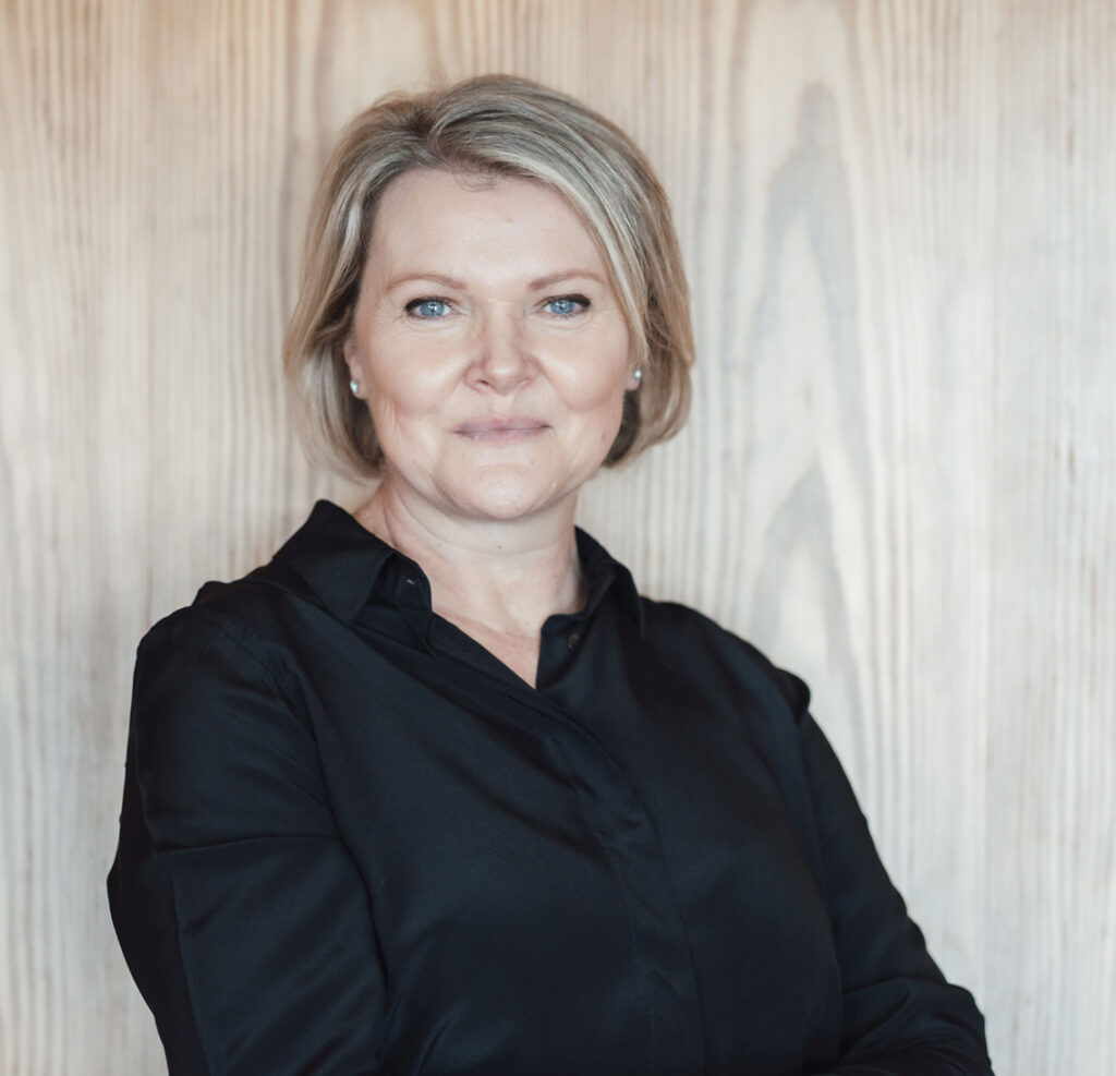 Porträtfoto einer Frau mittleren Alters vor hellem Holz­hintergrund. Sie steht frontal zur Kamera, die Arme locker vor dem Körper verschränkt, und blickt mit ruhigem, leicht lächelndem Ausdruck direkt in die Kamera. Sie hat kurzes, blondes Haar im Bob-Schnitt, helle Augen und trägt kleine Ohrstecker. Bekleidet ist sie mit einer schlichten, schwarzen Bluse mit V-Ausschnitt. Das Bild ist hell ausgeleuchtet und wirkt sachlich und professionell.