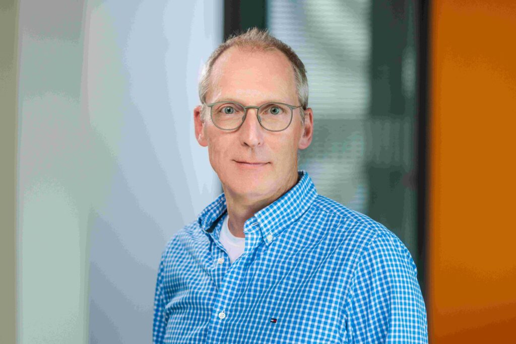 Ein Mann mittleren Alters steht frontal zur Kamera und blickt ruhig und aufmerksam in die Linse. Er hat kurzes, graumeliertes Haar, trägt eine runde Brille mit dünnem Metallrahmen und ein blau-weiß kariertes Hemd über einem hellen T-Shirt. Der Hintergrund ist unscharf und wirkt modern, mit hellen Flächen und einem vertikalen orangefarbenen Element auf der rechten Seite. Die Aufnahme ist gut ausgeleuchtet und vermittelt einen sachlichen, professionellen Eindruck.