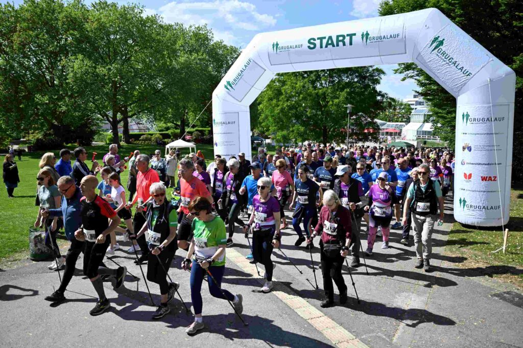 Eine große Gruppe von Teilnehmenden startet bei einem Lauf im Grugapark in Essen. Sie gehen und laufen unter einem weißen Startbogen mit der Aufschrift „GRUGALAUF START“ hindurch. Menschen unterschiedlichen Alters tragen Sportkleidung, viele haben Startnummern, einige nutzen Walkingstöcke. Die Stimmung wirkt lebhaft und gemeinschaftlich. Im Hintergrund sind grüne Bäume, Rasenflächen und Parkgebäude bei sonnigem Wetter zu sehen.