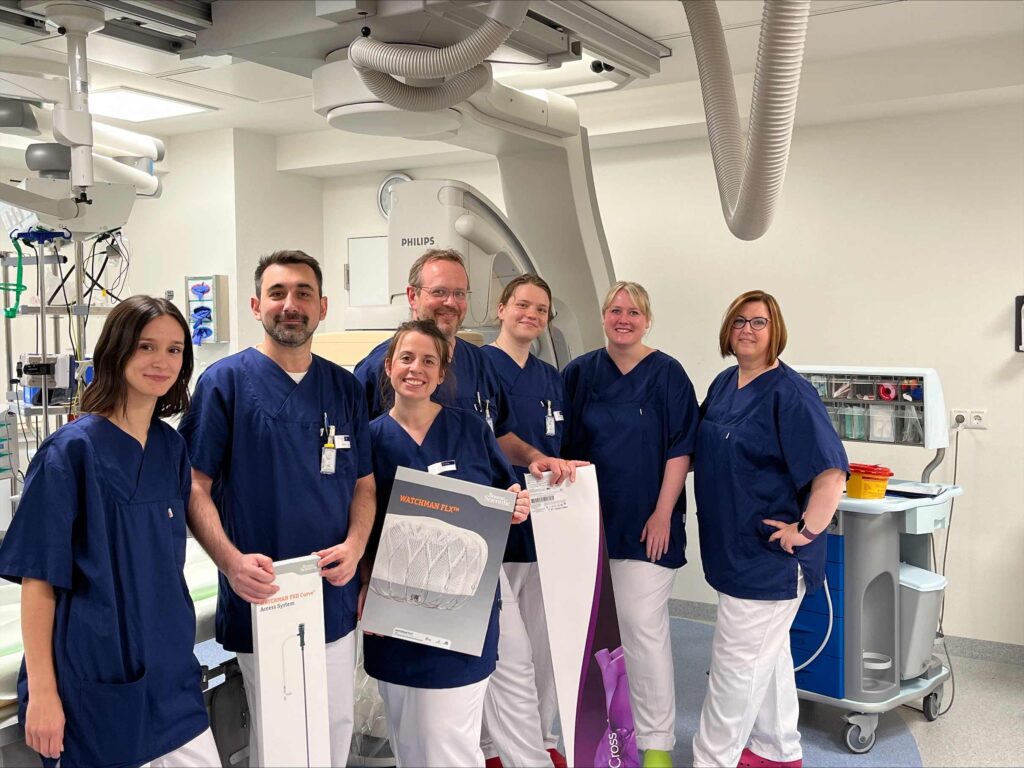 Sieben medizinische Fachkräfte stehen in einem hellen, modernen Eingriffsraum vor einem großen bildgebenden Medizingerät. Alle tragen dunkelblaue Kasacks und weiße Hosen. Die Gruppe blickt freundlich in die Kamera, einige lächeln. Zwei Personen halten Verpackungen von medizinischen Implantaten oder Instrumenten in den Händen. Im Hintergrund sind technische Geräte, Schläuche, Monitore und ein fahrbarer Gerätewagen zu sehen. Die Atmosphäre wirkt professionell und kollegial, der Raum ist sauber und klinisch eingerichtet.