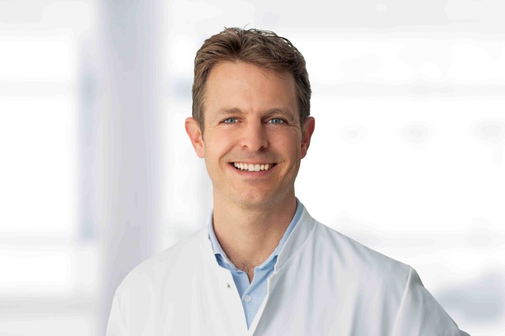 Porträt eines Mannes von der Brust aufwärts, der freundlich in die Kamera lächelt. Er hat kurze braune Haare, helle Augen und trägt einen weißen Arztkittel über einem hellblauen Hemd. Der Hintergrund ist hell und unscharf, mit weichen Lichtflächen, die an Fenster oder einen Klinikflur erinnern. Die Aufnahme wirkt professionell und ruhig, mit gleichmäßiger Beleuchtung und neutraler Farbgebung.