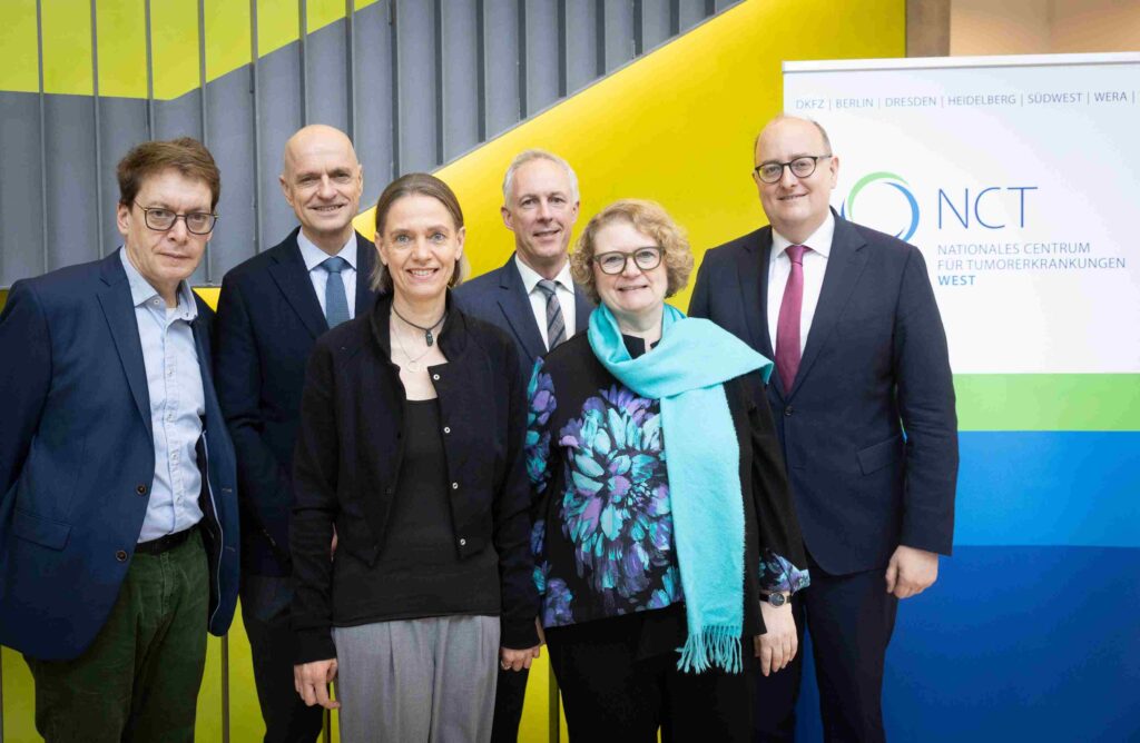 Sieben erwachsene Personen stehen dicht nebeneinander und blicken freundlich in die Kamera. Sie tragen überwiegend formelle Kleidung wie Anzüge, Sakkos und Blusen. Im Hintergrund ist eine moderne Wand mit schrägen Farbflächen in Gelb, Grau und Grün zu sehen. Rechts im Bild steht ein Roll-up mit dem Logo und der Aufschrift „NCT – Nationales Centrum für Tumorerkrankungen West“. Die Gruppe wirkt professionell und zugewandt; die Aufnahme vermittelt den Eindruck eines offiziellen Treffens oder Termins im wissenschaftlich-medizinischen Kontext.