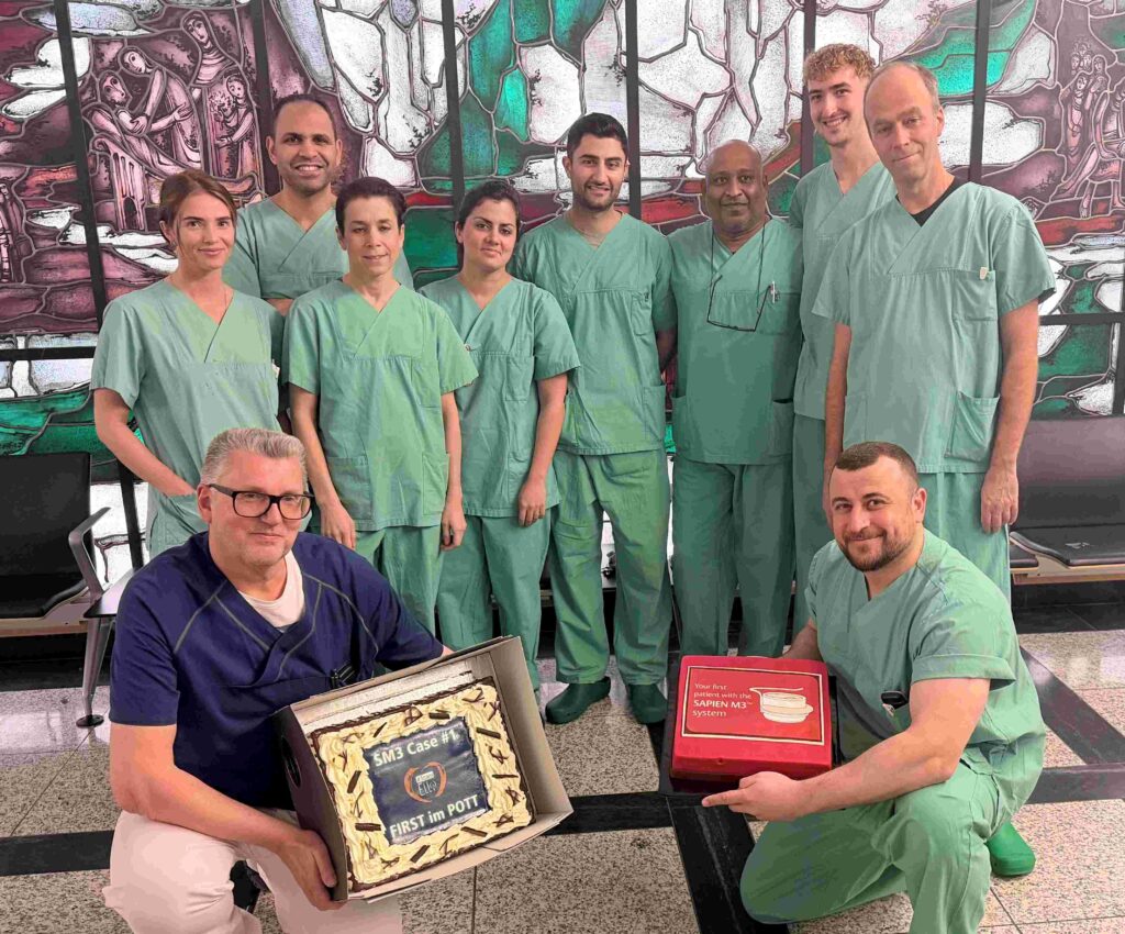 Gruppenfoto eines medizinischen Teams in einem Klinikfoyer. Neun Personen in grüner OP-Kleidung stehen nebeneinander vor einem großen, farbigen Glasfenster mit abstrakten und figürlichen Motiven in Grün-, Rot- und Weißtönen. Zwei weitere Männer befinden sich im Vordergrund: Links sitzt ein Mann mit grauem Haar und Brille in dunkelblauer Kasack-Oberbekleidung und heller Hose. Er hält eine geöffnete Schachtel mit einer rechteckigen Torte, auf der unter anderem „SM3 Case #1“ und „FIRST im POTT“ zu lesen ist. Rechts kniet ein Mann in grüner OP-Kleidung und hält eine rote Box mit der Aufschrift „Your first patient with the SAPIEN M3 system“. Die Gruppe blickt in die Kamera und wirkt stolz und zufrieden. Der Boden ist hell gefliest, seitlich stehen schwarze Sitzbänke.