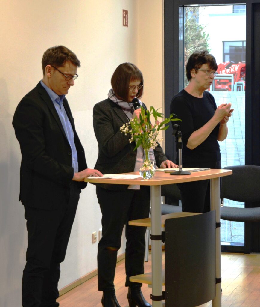 Drei Personen stehen nebeneinander an einem hohen Rednerpult in einem hellen Innenraum. In der Mitte spricht eine Person mit kurzem braunem Haar in ein Handmikrofon und liest dabei aus einem Blatt Papier. Links daneben steht eine weitere Person im dunklen Anzug, die ebenfalls auf Unterlagen blickt. Rechts steht eine Person in schwarzer Kleidung, die mit den Händen gestikulierend spricht; vor ihr befindet sich ein weiteres Mikrofon auf einem Stativ. Auf dem Rednerpult steht eine kleine Glasvase mit frischen Blumen. Im Hintergrund ist eine Glastür zu sehen, durch die Tageslicht einfällt und ein Außenbereich erkennbar ist. Die Szene wirkt wie ein formeller Programmpunkt bei einer Veranstaltung oder Zeremonie.