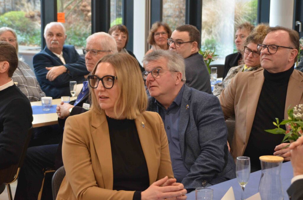 Mehrere erwachsene Personen sitzen dicht beieinander an Tischen in einem hellen Veranstaltungsraum und blicken aufmerksam nach vorne. Im Vordergrund sitzt eine Person mit schulterlangem blondem Haar und schwarzer Brille, bekleidet mit einem beigefarbenen Blazer über einem dunklen Oberteil. Neben und hinter ihr sitzen weitere Personen in formeller bis halbformeller Kleidung, teils mit Brillen. Auf den Tischen stehen Wassergläser, Sektgläser und Karaffen. Große Fenster im Hintergrund lassen Tageslicht herein und geben den Blick auf Bäume und Grünflächen frei. Die Atmosphäre wirkt konzentriert und ruhig, wie bei einem Vortrag oder einer offiziellen Veranstaltung.