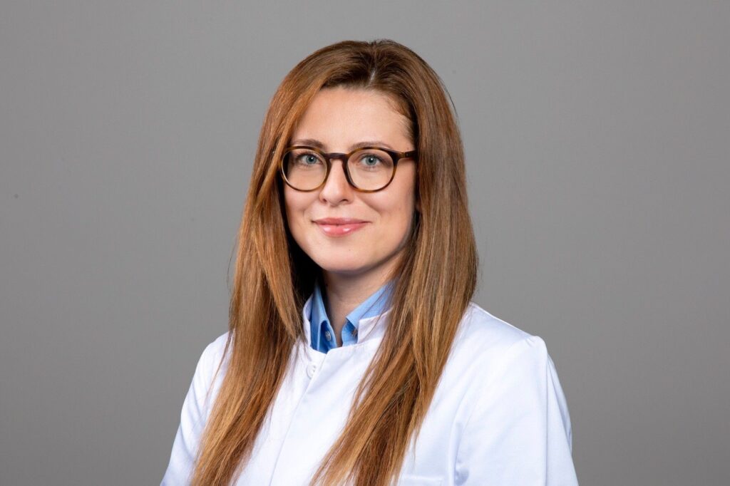 orträt einer Frau mit langen, glatten, hellbraunen Haaren, die seitlich gescheitelt sind. Sie trägt eine runde Brille mit dunklem Rahmen und blickt ruhig und freundlich in die Kamera. Bekleidet ist sie mit einem weißen Arztkittel über einer hellblauen Bluse. Der Hintergrund ist neutral grau, die Aufnahme wirkt sachlich und professionell.