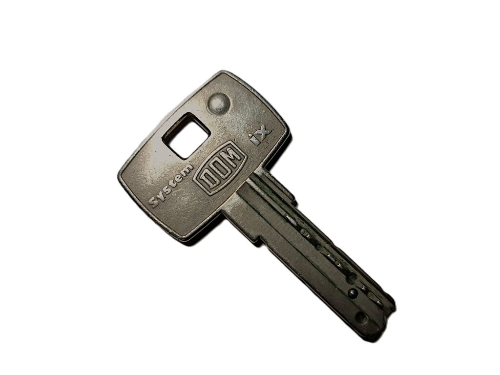 „Metallischer Euroschlüssel mit rechteckigem Kopf und kurzem Schaft, schräg vor hellem Hintergrund fotografiert. Auf dem Schlüsselkopf ist die Prägung ‚System DOM‘ zu sehen sowie eine kleine quadratische Öffnung.