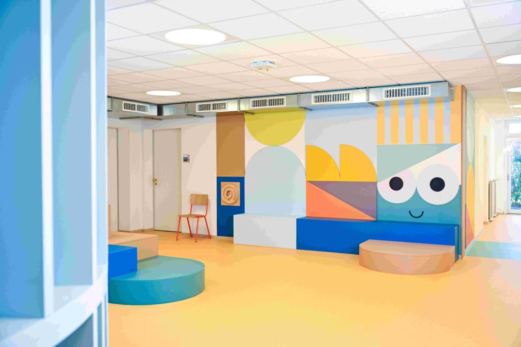 Heller, freundlich gestalteter Innenraum, vermutlich ein Warte- oder Aufenthaltsbereich in einer Kinder- oder Therapieeinrichtung. Der Boden ist in warmem Gelb gehalten, die Wände sind mit großen, bunten geometrischen Formen in Blau, Grün, Gelb, Orange und Beige bemalt. Rechts an der Wand ist ein verspieltes Gesicht mit zwei großen runden Augen und einem kleinen lächelnden Mund zu sehen. Entlang der Wände stehen niedrige, gepolsterte Sitzbänke in Blau und Beige sowie runde Podeste in Grün und Holzoptik. Links befinden sich zwei geschlossene weiße Türen, daneben ein einzelner Holzstuhl mit rotem Metallgestell. Die Decke ist mit weißen Akustikplatten und runden Deckenleuchten ausgestattet, darüber verlaufen sichtbare Lüftungsschächte. Am Ende des Flurs fällt Tageslicht durch eine Glastür oder ein Fenster ein.