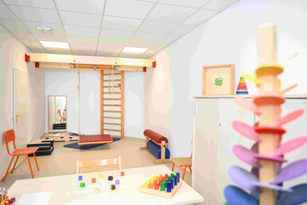 Heller Therapieraum mit kindgerechter Ausstattung. Im Hintergrund ist eine große Holzkonstruktion an der Wand befestigt, bestehend aus einer Sprossenwand und einem Querbalken mit Seilen. Daran hängt eine rechteckige Schaukel mit rotem Polster über einer blauen Matte. Rechts daneben steht eine schmale Turnbank mit roter Auflage, darunter liegen blaue Rollen. Links im Raum stehen zwei kleine orangefarbene Stühle. An der linken Wand befindet sich ein großer Spiegel, vor dem mehrere schwarze Trittstufen liegen. Im Vordergrund steht ein niedriger weißer Tisch mit kleinen bunten Holzklötzen und einem Steckspiel in Regenbogenfarben. Rechts vorne, unscharf im Bild, ist ein vertikales Holzspielzeug mit bunten Scheiben zu sehen. An der rechten Wand steht ein hoher weißer Schrank; darauf liegen ein Stapelturm aus bunten Ringen und ein gerahmtes Bild mit einem grünen Kleeblatt. Der Raum wirkt aufgeräumt, freundlich und auf Bewegungs- und Spieltherapie ausgerichtet.