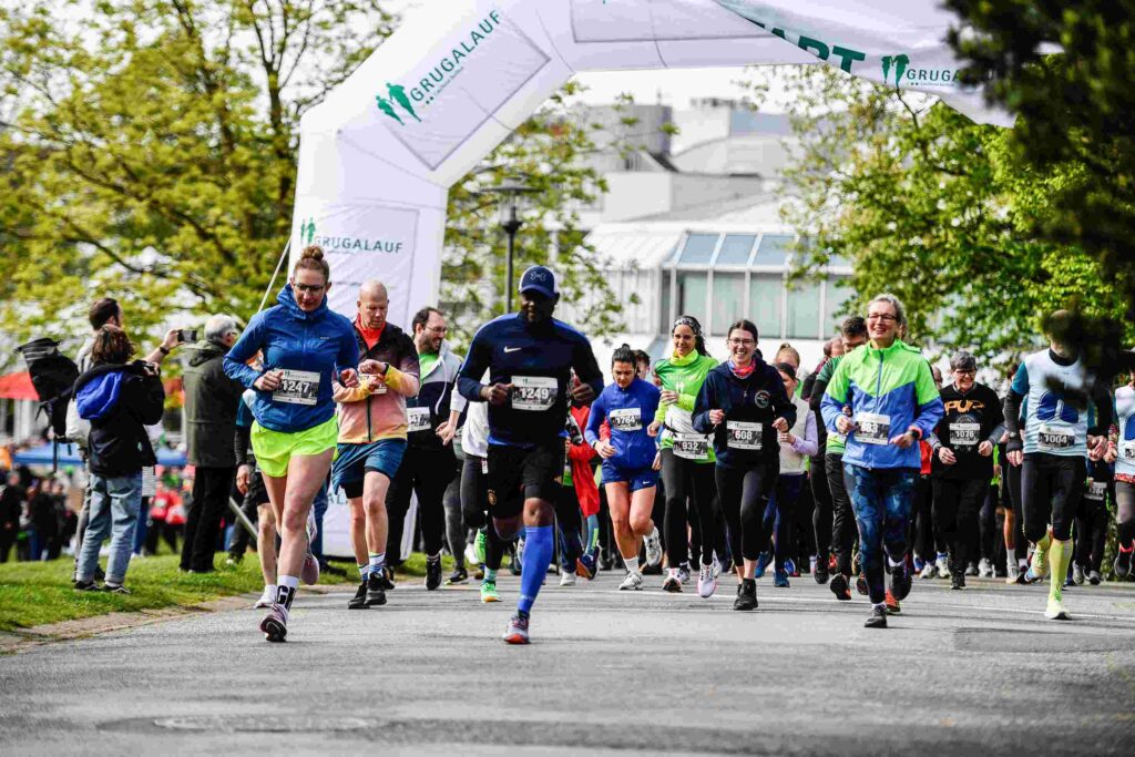 Eine große Gruppe von Läuferinnen und Läufern startet gemeinsam unter einem weißen Startbogen mit der Aufschrift „GRUGALAUF“. Im Vordergrund laufen mehrere Personen nebeneinander auf einer asphaltierten Strecke durch eine grüne Parkanlage. Sie tragen Sportkleidung in kräftigen Farben wie Blau, Grün und Schwarz sowie Startnummern auf der Brust. Links vorne läuft eine Frau mit blauem Oberteil und neongrüner Shorts, in der Mitte ein Mann in dunkelblauer Laufkleidung mit Kappe, rechts eine Frau mit blau-grüner Jacke. Im Hintergrund stehen weitere Teilnehmende und Zuschauende zwischen Bäumen; dahinter sind helle Gebäude zu sehen. Die Stimmung wirkt energiegeladen und gemeinschaftlich.