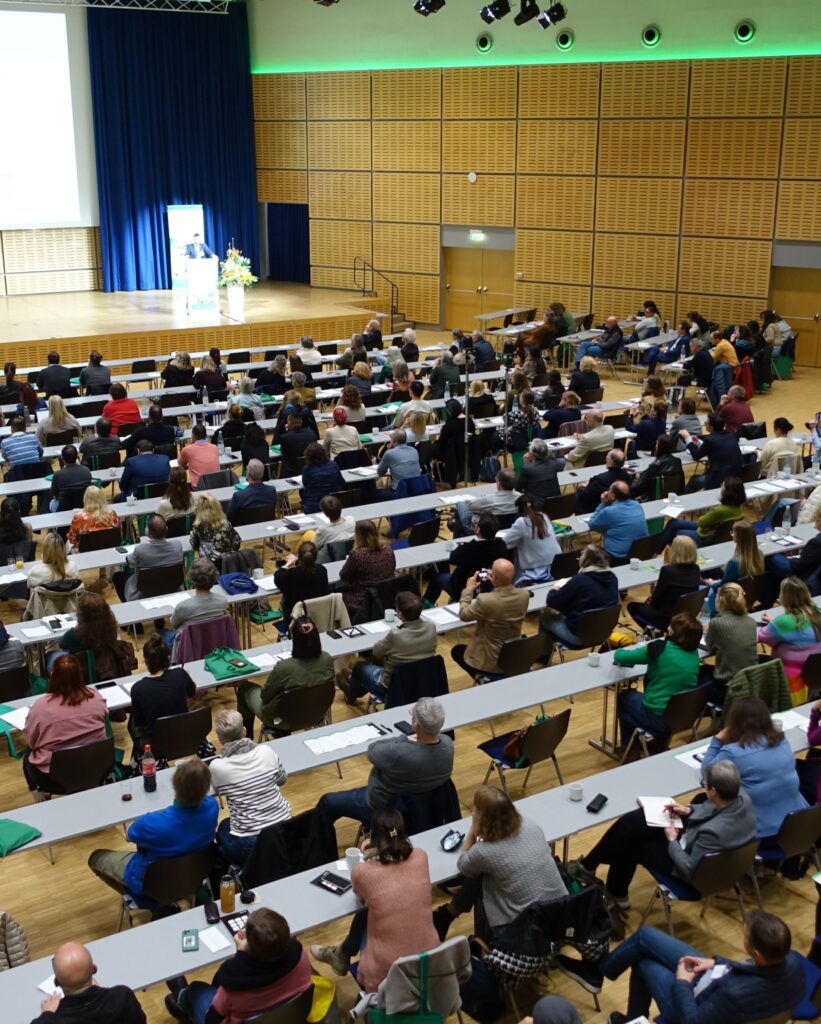 Großer Hörsaal mit ansteigenden Sitzreihen, in dem zahlreiche Erwachsene dicht nebeneinander an langen Tischen sitzen und nach vorne blicken. Viele haben Notizblöcke, Getränke oder Taschen vor sich. Links vorne steht eine Person an einem Rednerpult auf einer Bühne neben einer großen Projektionsfläche und einem Blumenarrangement. Die Rückwand ist mit hellen Holzpaneelen verkleidet, darüber verläuft eine grüne Lichtleiste; Scheinwerfer hängen an der Decke. Die Atmosphäre wirkt wie eine gut besuchte Fachveranstaltung oder ein Symposium.