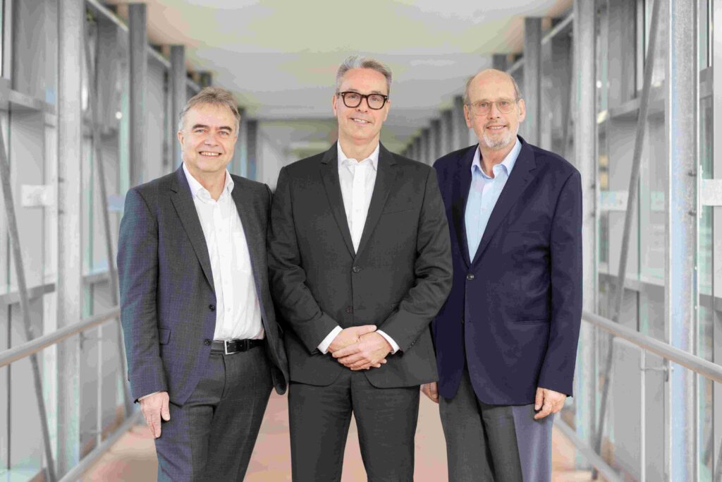 Drei Männer stehen nebeneinander in einem hellen, verglasten Verbindungsgang und blicken in die Kamera. Alle tragen Anzüge mit hellen Hemden; der Mann in der Mitte trägt zusätzlich eine dunkle Brille und hat die Hände vor dem Körper gefaltet. Die beiden Männer links und rechts stehen mit entspannten Armen und leichtem Lächeln. Der Hintergrund zeigt eine moderne Architektur mit großen Fensterflächen, Metallstreben und einem durchgehenden Handlauf entlang des Gangs.