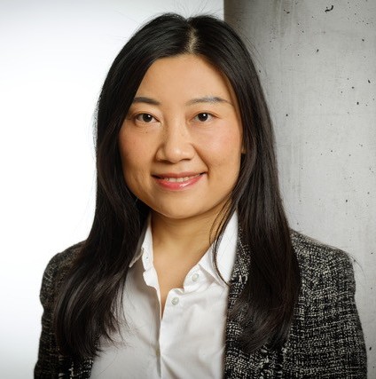 Porträt einer Frau mit langem, glattem, schwarzem Haar, das offen über die Schultern fällt. Sie blickt direkt in die Kamera und lächelt freundlich. Sie trägt eine weiße Bluse mit offenem Kragen und darüber ein dunkel gemustertes Sakko. Der Hintergrund ist neutral gehalten: links hell, rechts eine graue Betonwand. Die Aufnahme ist als klassisches Business-Porträt im Hochformat gestaltet.