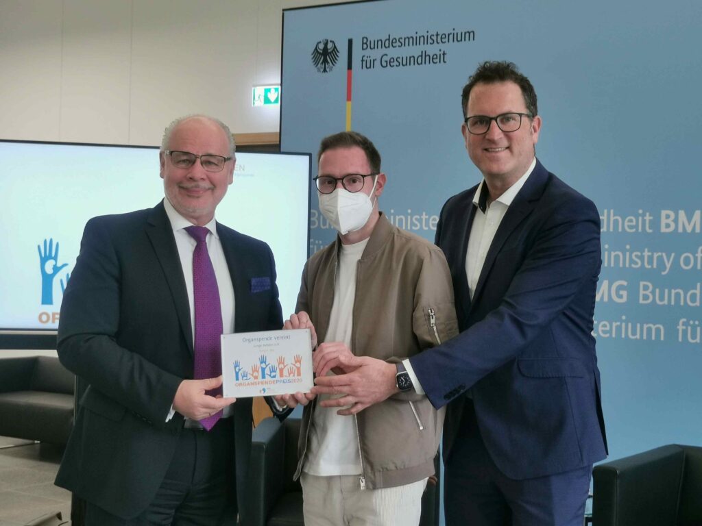 Drei Männer stehen nebeneinander in einem hellen Besprechungsraum und halten gemeinsam eine Urkunde in die Kamera. Links ein älterer Mann im dunklen Anzug mit Krawatte, rechts ein weiterer Mann im Anzug; beide lächeln. In der Mitte steht ein jüngerer Mann mit Brille und weißer FFP2-Maske, leger gekleidet mit heller Jacke. Auf der Urkunde sind bunte Hände abgebildet und der Schriftzug „Organspendepreis 2026“ erkennbar. Im Hintergrund ist eine Wand mit dem Logo und Schriftzug des Bundesministeriums für Gesundheit zu sehen.