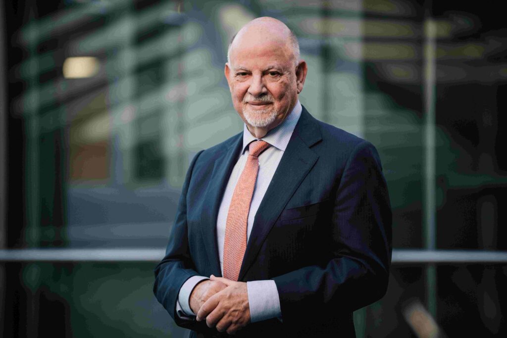 Porträt eines älteren Mannes mit Glatze und kurzem weißem Bart. Er trägt einen dunklen Anzug, ein helles Hemd und eine orangefarbene Krawatte und steht leicht seitlich zur Kamera, die Hände locker vor dem Körper gefaltet. Sein Gesicht zeigt einen ruhigen, freundlichen Ausdruck. Im Hintergrund ist unscharf eine moderne Glas- und Stahlarchitektur zu sehen, vermutlich ein Innen- oder Eingangsbereich eines größeren Gebäudes.