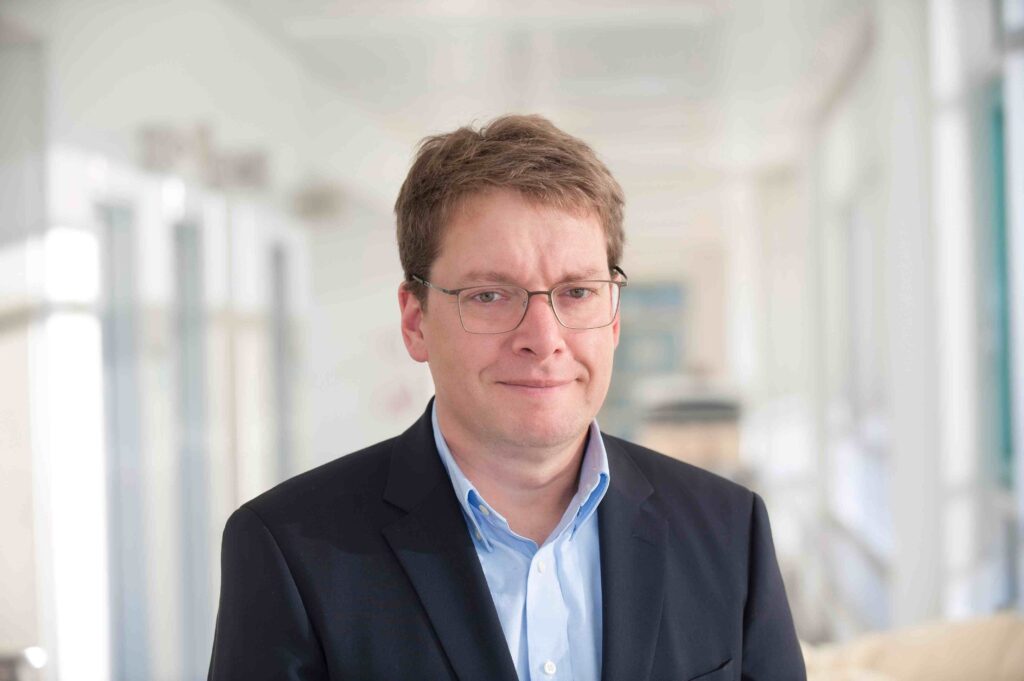 Porträt eines Mannes mittleren Alters mit kurzen braunen Haaren und rechteckiger Brille. Er trägt ein hellblaues Hemd und ein dunkles Sakko und blickt mit leichtem, neutralem Lächeln direkt in die Kamera. Die Aufnahme ist von der Brust aufwärts fotografiert. Im Hintergrund ist unscharf ein heller Krankenhaus- oder Laborflur mit weißen Wänden und medizinischen Geräten zu erkennen.