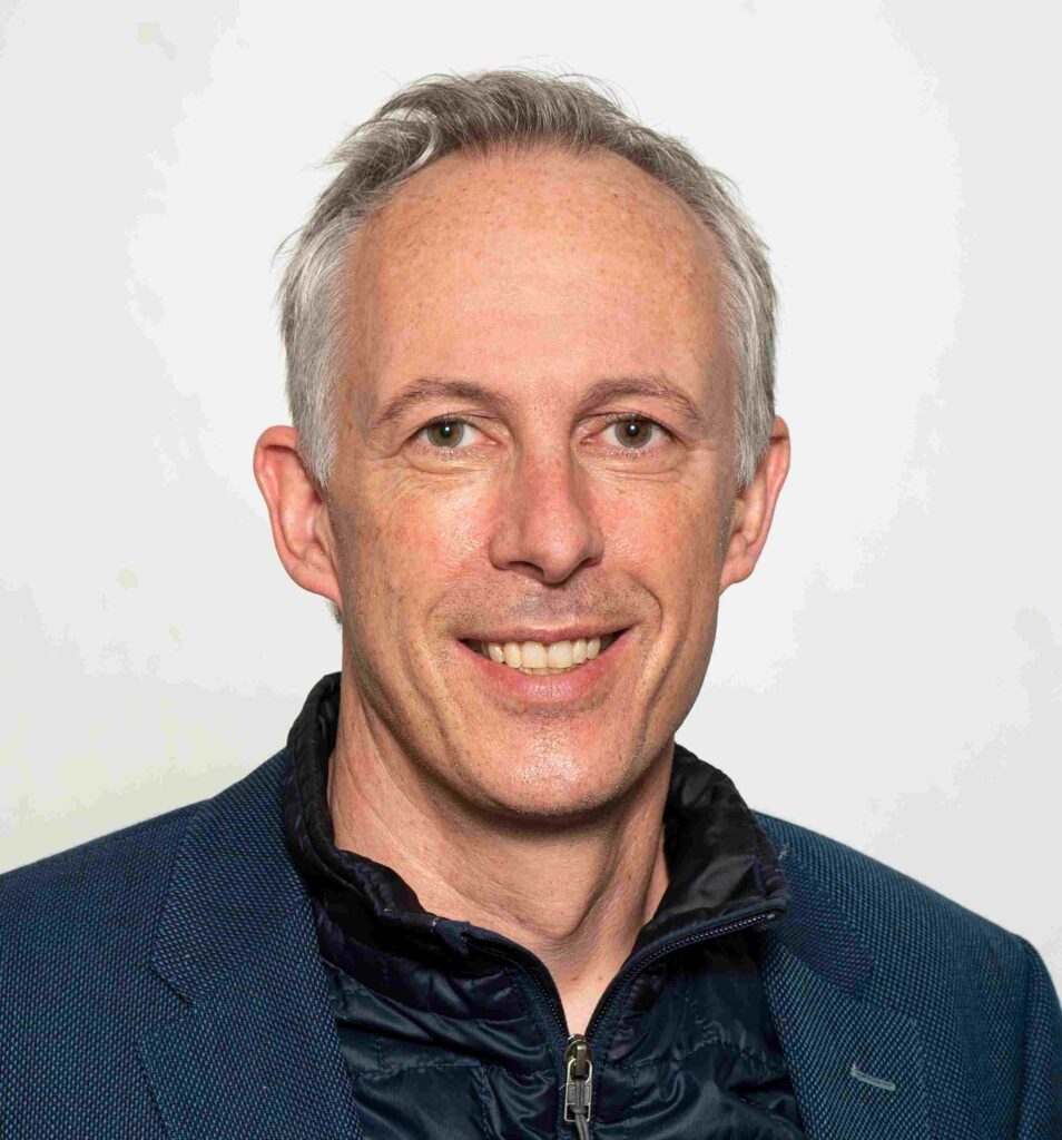 Porträt eines lächelnden Mannes mittleren Alters mit kurzen grauen Haaren und heller Haut vor neutralem, hellgrauem Hintergrund. Er blickt direkt in die Kamera und trägt ein dunkelblaues Sakko über einer schwarzen, leicht glänzenden Steppjacke mit Reißverschluss.