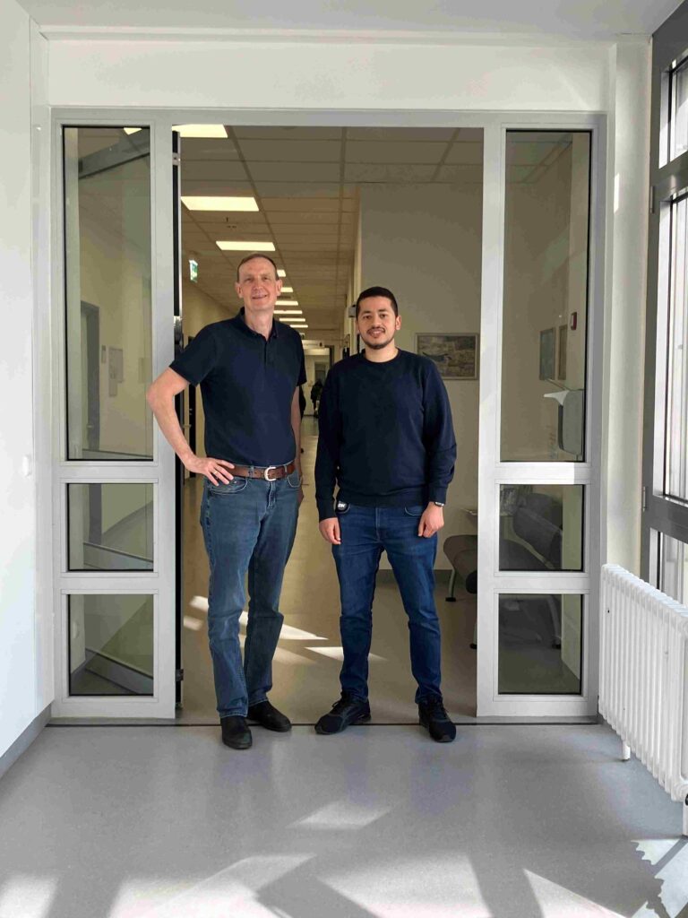 Zwei Männer stehen nebeneinander in einer offenen Glastür in einem hellen Flur, vermutlich in einem Büro- oder Klinikgebäude. Beide blicken in die Kamera und lächeln. Der Mann links ist größer, trägt ein dunkles Poloshirt, Jeans und einen Gürtel, eine Hand lässig in die Hüfte gestützt. Der Mann rechts ist etwas kleiner, trägt einen dunklen Pullover und Jeans, die Arme locker an den Seiten. Hinter ihnen erstreckt sich ein langer, gut beleuchteter Gang mit Deckenleuchten und weiteren Türen. Rechts im Vordergrund ist ein Fenster mit Heizkörper darunter, durch das Tageslicht einfällt.