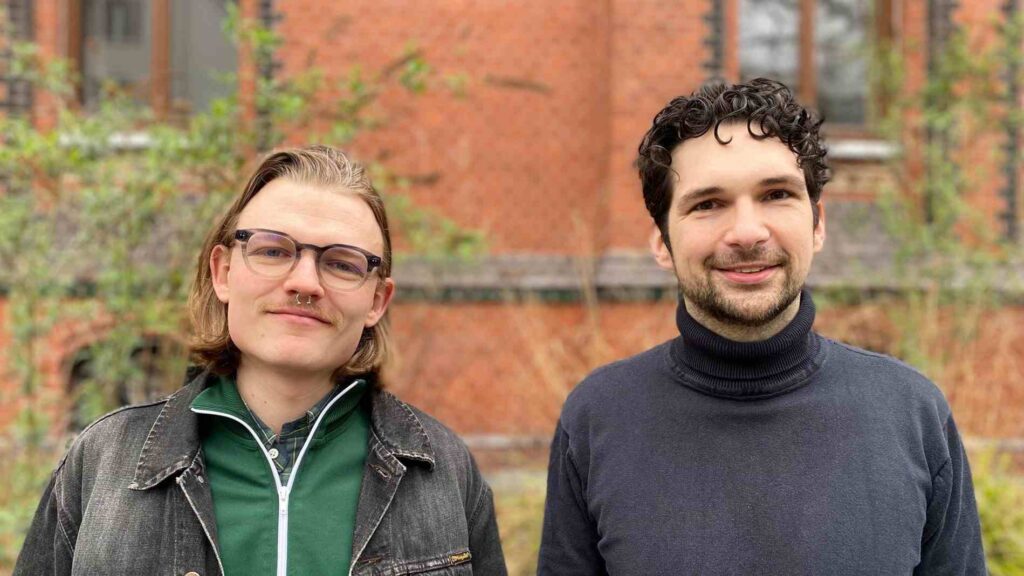 Zwei Männer stehen nebeneinander im Freien und blicken in die Kamera. Links eine Person mit schulterlangen, hellbraunen Haaren, Brille und kleinem Schnurrbart sowie Nasenpiercing; sie trägt eine grüne Jacke unter einer dunklen Jeansjacke. Rechts eine Person mit kurzen, dunklen Locken und leichtem Bart, bekleidet mit einem dunklen Rollkragenpullover. Beide lächeln leicht. Im Hintergrund ist unscharf ein rötliches Backsteingebäude mit Fenstern und etwas Bewuchs zu erkennen.
