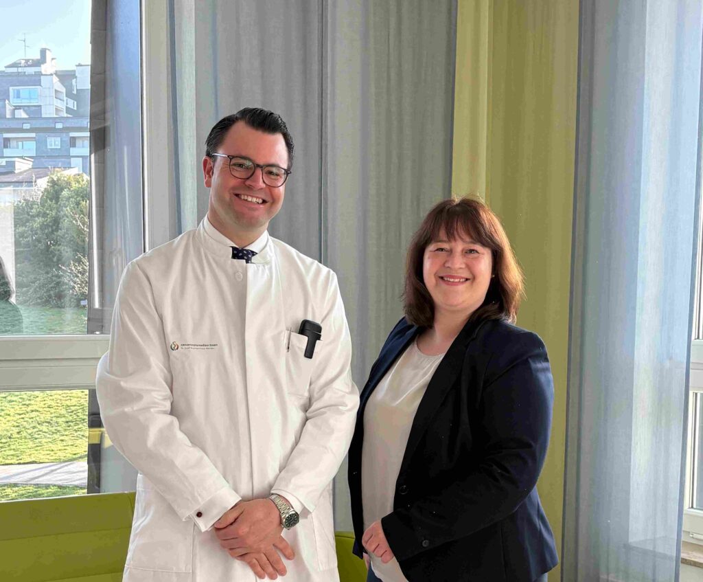Zwei Personen stehen nebeneinander vor großen Fenstern in einem hellen Raum. Links ein Mann in weißem Arztkittel mit Brille und dunklem Haar, die Hände locker vor dem Körper gefaltet; in der Brusttasche steckt ein schwarzes Gerät. Rechts eine Frau mit schulterlangem braunem Haar, die ein dunkles Sakko über einer hellen Bluse trägt. Beide lächeln in die Kamera. Im Hintergrund sind Vorhänge in gedeckten Farben sowie draußen Gebäude und Grünflächen zu erkennen.