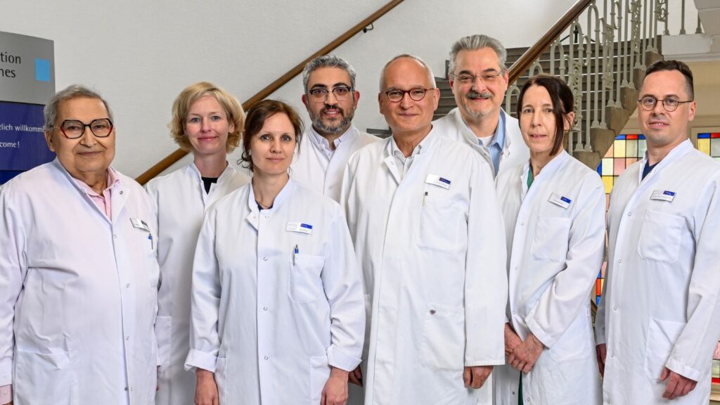 Sieben medizinische Fachkräfte in weißen Arztkitteln stehen gemeinsam für ein Gruppenfoto in einem hellen Treppenhaus einer Klinik oder medizinischen Einrichtung. Die Gruppe besteht aus Frauen und Männern unterschiedlichen Alters. Einige tragen Brillen, alle blicken freundlich oder neutral in die Kamera. Im Hintergrund sind eine geschwungene Treppe mit dekorativem Geländer sowie farbige Glaselemente zu sehen. Die Aufnahme vermittelt einen professionellen und kollegialen Eindruck eines interdisziplinären Teams im Bereich der Gefäßchirurgie oder Medizin.
