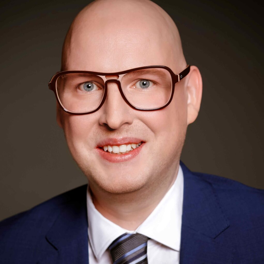 Porträtaufnahme eines lächelnden Mannes mit heller Haut und kahlem Kopf vor dunklem Hintergrund. Er trägt eine große, dunkelbraune Brille mit leicht transparentem Rahmen, ein weißes Hemd, eine gestreifte Krawatte in Blau- und Grautönen sowie ein dunkelblaues Sakko. Die Person blickt direkt in die Kamera und wirkt freundlich und professionell. Das Bild ist als klassisches Business-Porträt mit weicher Ausleuchtung aufgenommen.