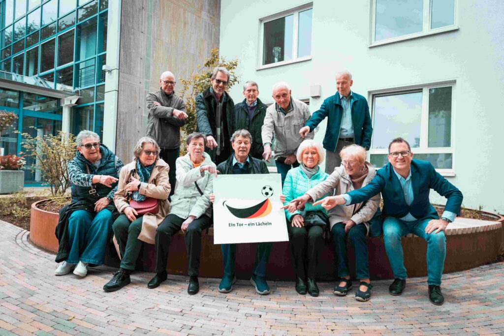 Eine Gruppe älterer Frauen und Männer sitzt und steht gemeinsam vor einem modernen Gebäude im Außenbereich. In der Mitte halten mehrere Personen ein Schild mit dem Logo und Schriftzug „Ein Tor – ein Lächeln“. Fast alle Beteiligten zeigen lächelnd mit ausgestrecktem Arm auf das Schild. Rechts außen kniet ein Mann im blauen Sakko vor der Gruppe. Die Szene vermittelt Gemeinschaft, Engagement und Unterstützung für das Projekt.