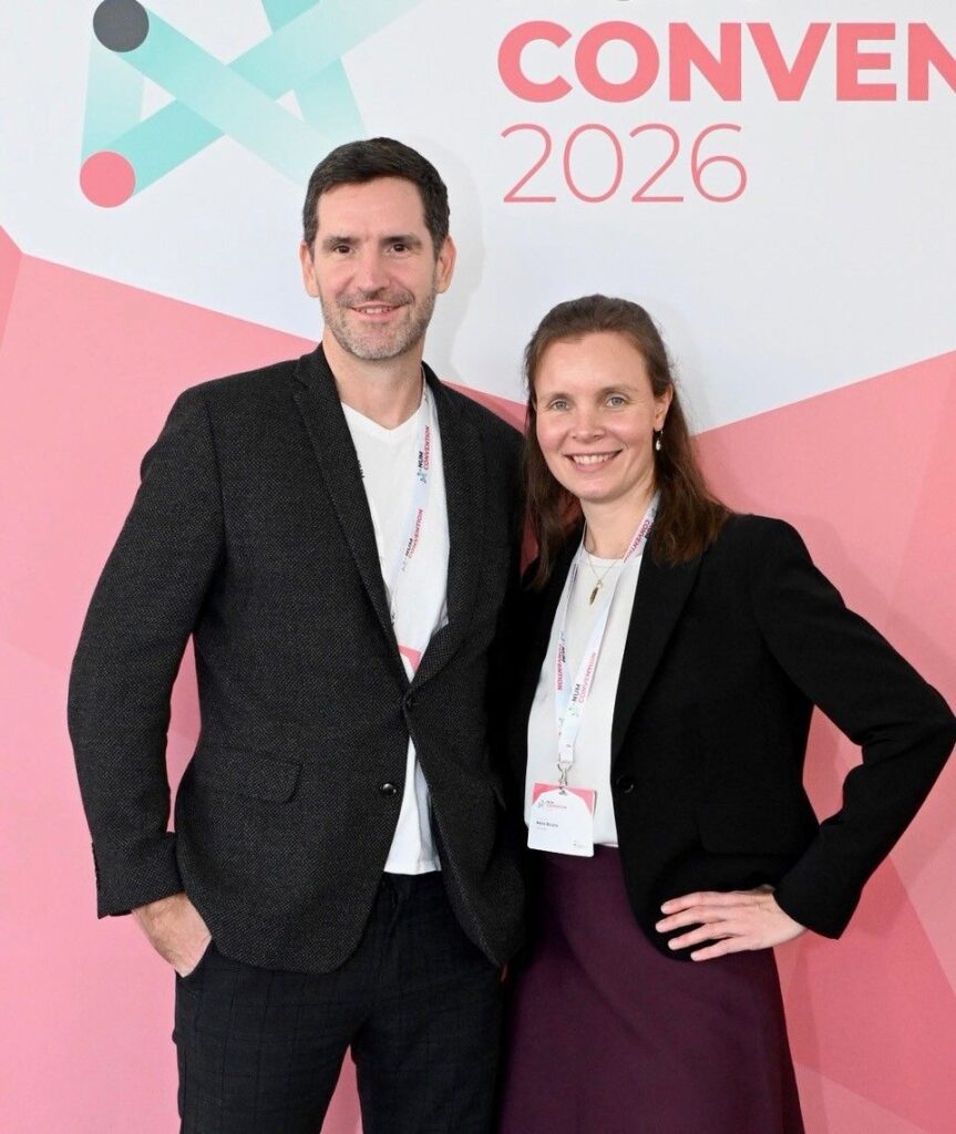 Zwei Personen stehen nebeneinander vor einer hell gestalteten Messewand mit grafischen Linien und der Aufschrift „CONVENT 2026“. Links ein Mann mit kurzen dunklen Haaren und leichtem Bart, gekleidet in ein dunkles Sakko über einem hellen T-Shirt; eine Hand steckt locker in der Hosentasche. Rechts eine Frau mit braunen, zurückgebundenen Haaren, die ein schwarzes Sakko, eine helle Bluse und einen dunklen Rock trägt; eine Hand liegt selbstbewusst in der Hüfte. Beide tragen Namensschilder an Umhängebändern und lächeln in die Kamera. Die Atmosphäre wirkt professionell und freundlich, typisch für eine Konferenz oder Branchenveranstaltung.
